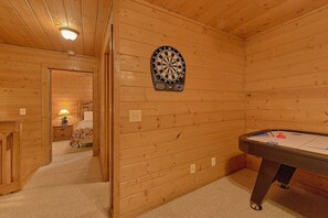 Une pièce aux murs en bois, avec un jeu de fléchettes, une table de billard et un lit doté d’une table de chevet.