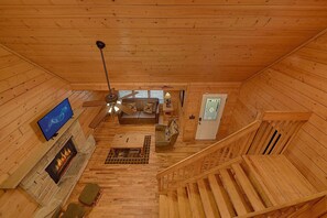 Interior de una cabaña de madera con chimenea, un sofá, un televisor y una escalera.