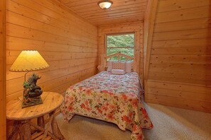 Une petite pièce chaleureuse aux murs en bois, avec un lit simple recouvert d’une literie à motifs floraux, une petite table avec une lampe et une vue sur la verdure à l’extérieur.