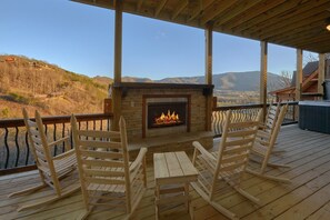 A wooden deck with a stone fireplace, wooden chairs, and a view of a mountainous landscape. - Hideaway Dreams | Theater, Smoky Mtn Views, Hot Tub, Game Room, Sleeps 12 + Free Attraction Tickets (Wears Valley)
