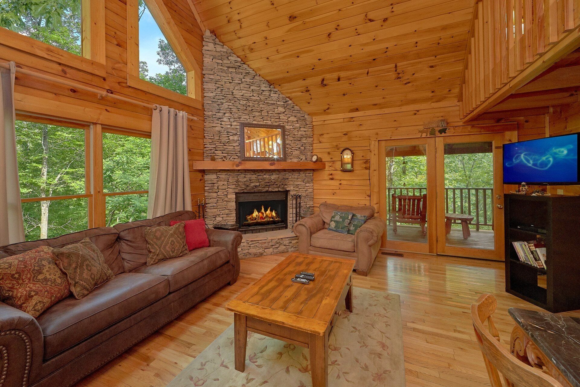 A cozy cabin interior with a stone fireplace, leather sofas, a wooden coffee table, and a television.