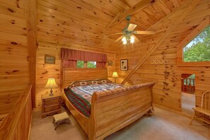A wooden cabin bedroom with a bed, nightstands, a lamp, a ceiling fan, and a window. - Jasmine's Retreat | Hot Tub, Game Room, Fireplace + Free Attraction Tickets (Wears Valley)