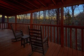 A wooden deck with two rocking chairs and a table, overlooking a forested area. - Just Barely Making It | Hot Tub, Fireplace + Free Attraction Tickets (Wears Valley)