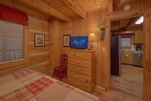 A wooden interior with a bed, a television, a chair, and a kitchen area. - Just Barely Making It | Hot Tub, Fireplace + Free Attraction Tickets (Wears Valley)