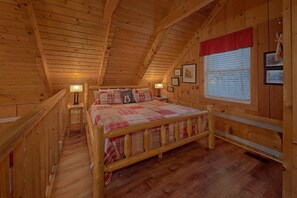 A cozy wooden cabin bedroom with a bed, two bedside lamps, a window with blinds, and framed pictures on the wall. - Just Barely Making It | Hot Tub, Fireplace + Free Attraction Tickets (Wears Valley)