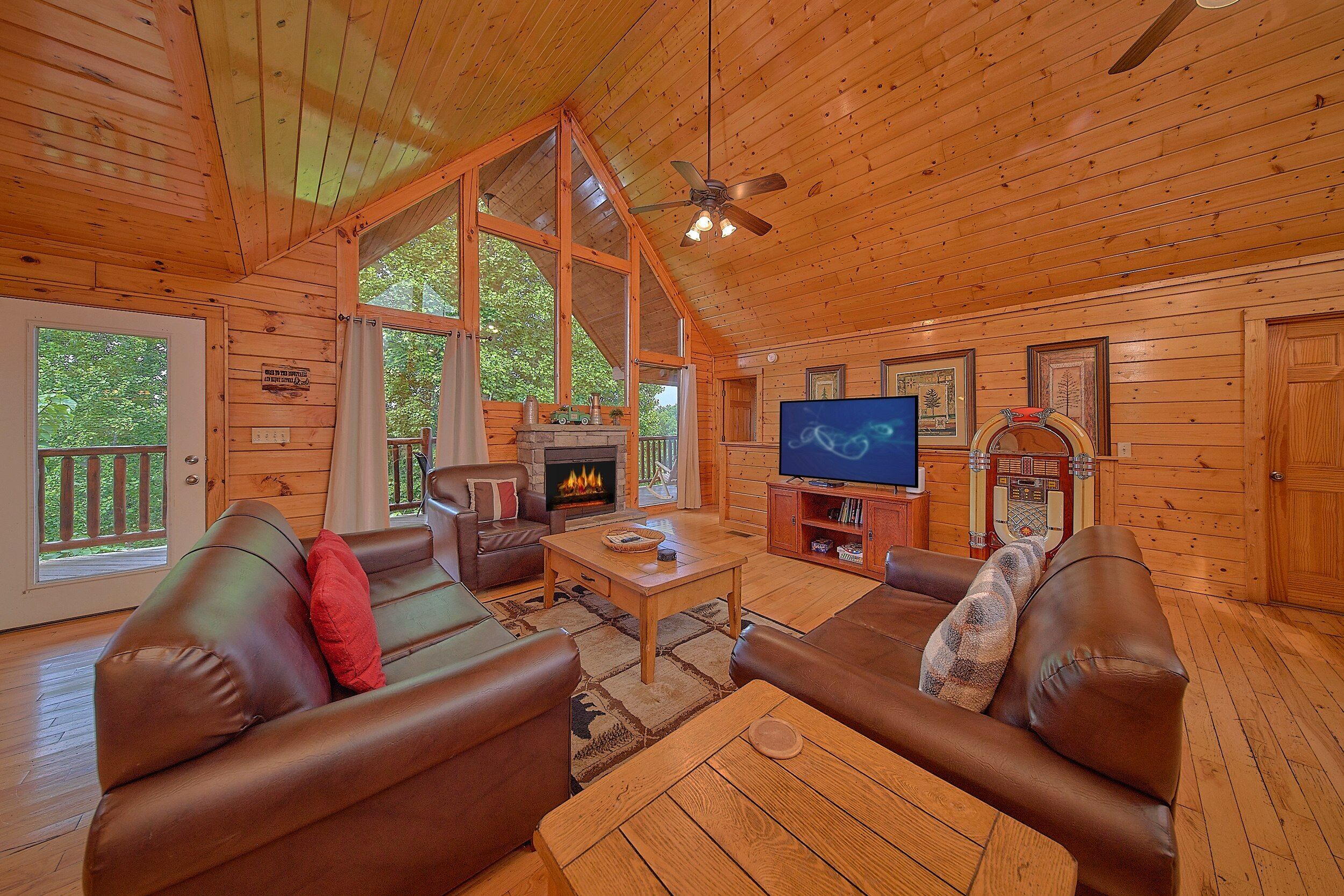 L’intérieur d’un chalet accueillant, avec une cheminée, des canapés en cuir, un téléviseur et une grande fenêtre donnant sur un paysage verdoyant.