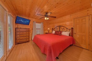 Ein Zimmer mit Holzwänden, einem Bett, einem Schminktisch, einem Fernseher und einem Deckenventilator.
