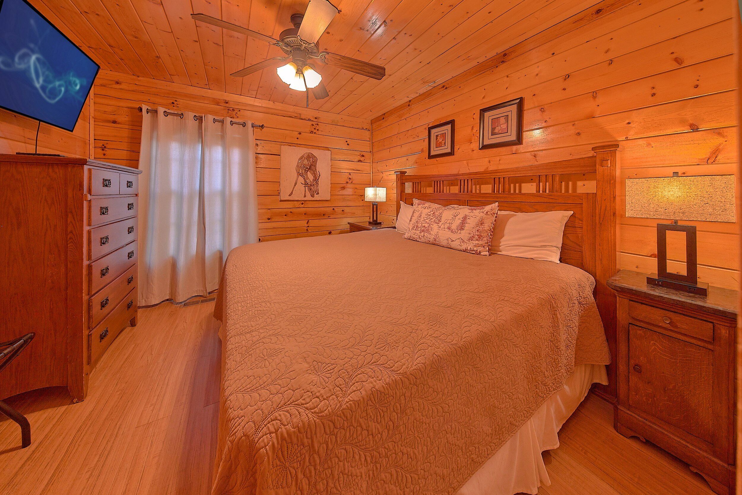 A wooden-paneled bedroom with a bed, nightstands, a dresser, a ceiling fan, and a television.