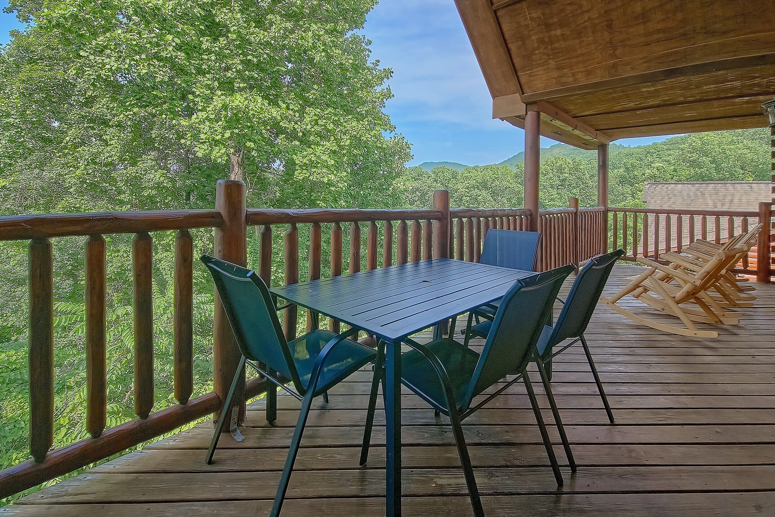 Eine Holzterrasse mit Tisch und Stühlen, umgeben von einem Holzgeländer, mit Blick auf ein bewaldetes Gebiet.