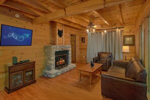 Interior acogedor de una cabaña de madera con chimenea de piedra, mobiliario de cuero y televisor montado en la pared.