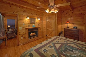 A cozy log cabin room with a fireplace, wooden walls, a ceiling fan, and a bed with a patterned bedspread. - Lazy View Lodge | Smoky Mtn Views, Hot Tub, Game Room, Fire Pit, Fireplace + Free Attraction Tickets (Sevierville)