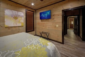 Ein Hotelzimmer mit holzgetäfelten Wänden, einem Bett mit floralem Überwurf, einem an der Wand montierten Fernseher und einem Flur, der in ein weiteres Zimmer führt.