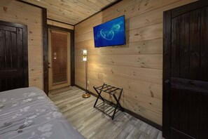 A room with wooden walls, a bed with a floral bedspread, a television mounted on the wall, and a small table with a ladder. - LeConte Views | Theater, Smoky Mtn Views, Hot Tub, Game Room, Sleeps 14 + Free Attraction Tickets (Gatlinburg)