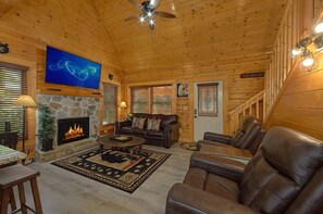 A cozy cabin interior with a stone fireplace, leather furniture, a flat-screen TV, and a staircase. - Lovers Paradise | Hot Tub, Game Room, Pigeon Forge, Resort Pool, Fireplace + Free Attraction Tickets (Pigeon Forge)