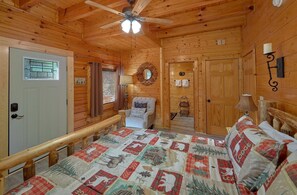 A log cabin bedroom with a bed, a chair, a ceiling fan, a window with curtains, a door, and a wall-mounted lamp. - Lovers Paradise | Hot Tub, Game Room, Pigeon Forge, Resort Pool, Fireplace + Free Attraction Tickets (Pigeon Forge)
