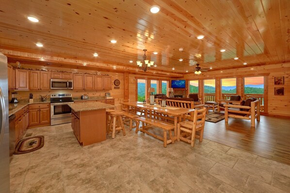 A spacious log cabin interior with a kitchen, dining area, and a large window overlooking a mountainous landscape.