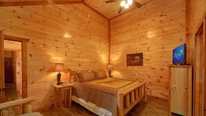A wooden cabin interior with a bed, nightstands, a television, and a ceiling fan.