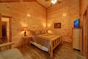 A wooden cabin interior with a bed, nightstands, a television, and a ceiling fan.