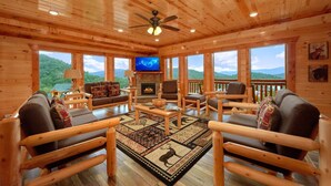 A log cabin interior with a fireplace, wooden furniture, and large windows offering mountain views.
