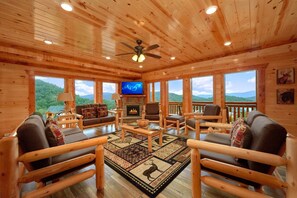 A log cabin interior with a fireplace, wooden furniture, and large windows offering mountain views.