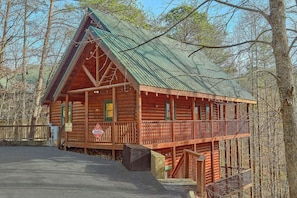 A log cabin with a porch and a metal roof, surrounded by trees. - Major Oaks | Theater, Hot Tub, Game Room, Sleeps 12, King Suites + Free Attraction Tickets (Pigeon Forge)