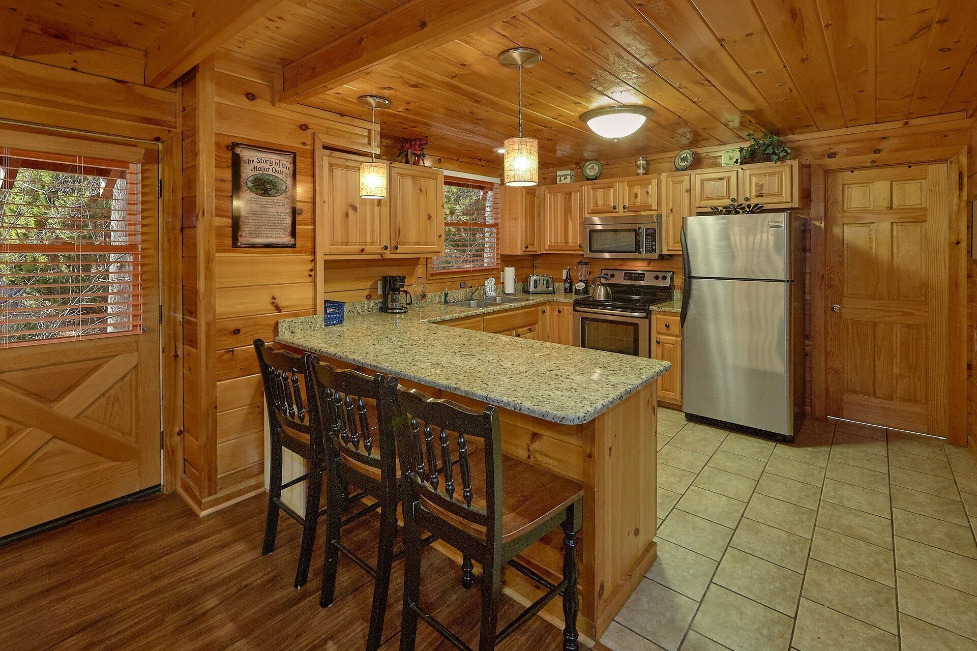 A wooden cabin interior with a kitchen featuring a granite countertop, wooden cabinets, a stainless steel refrigerator, and a bar area with chairs.