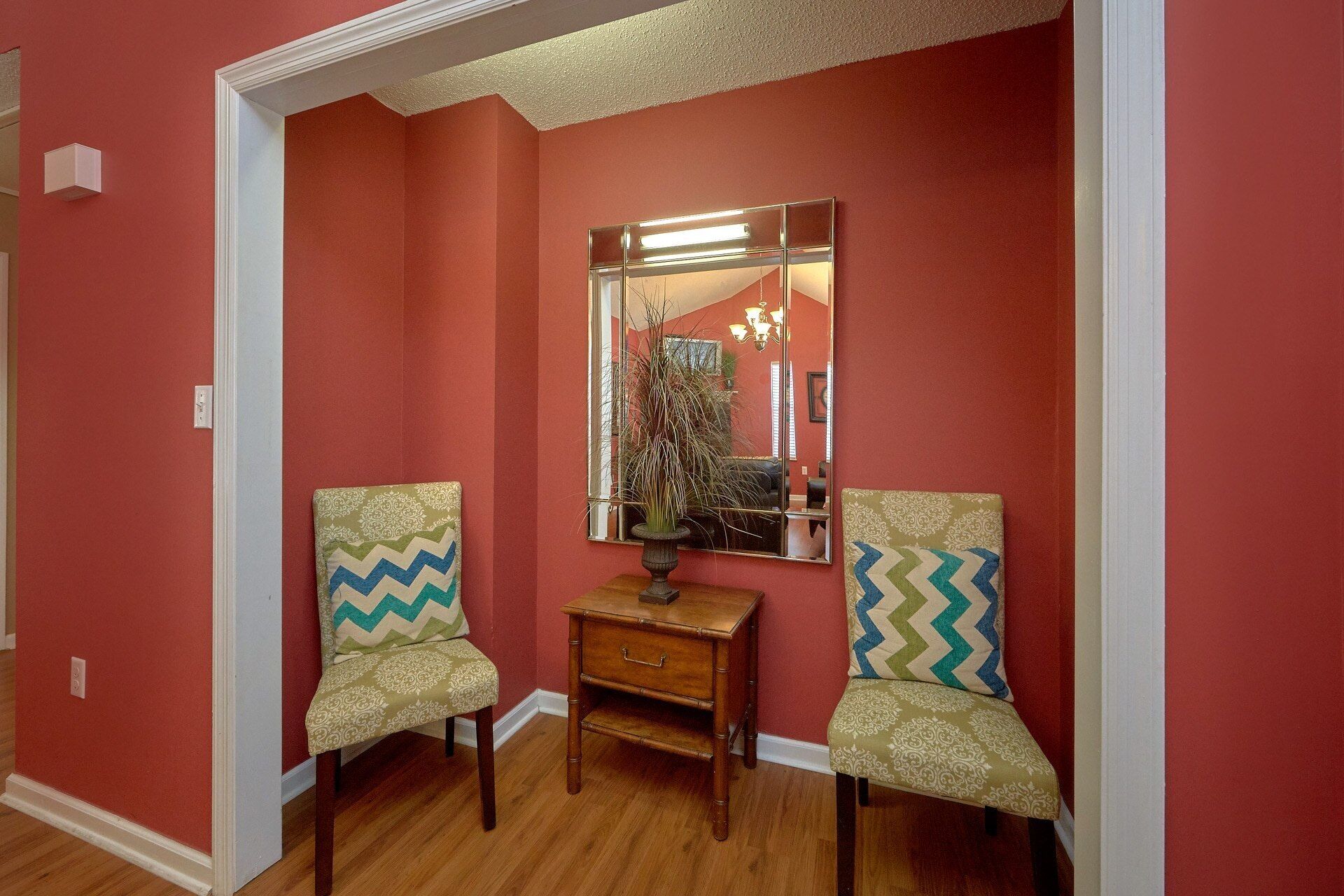 A hallway with red walls, two chairs with patterned cushions, a wooden side table, and a mirror reflecting a chandelier.