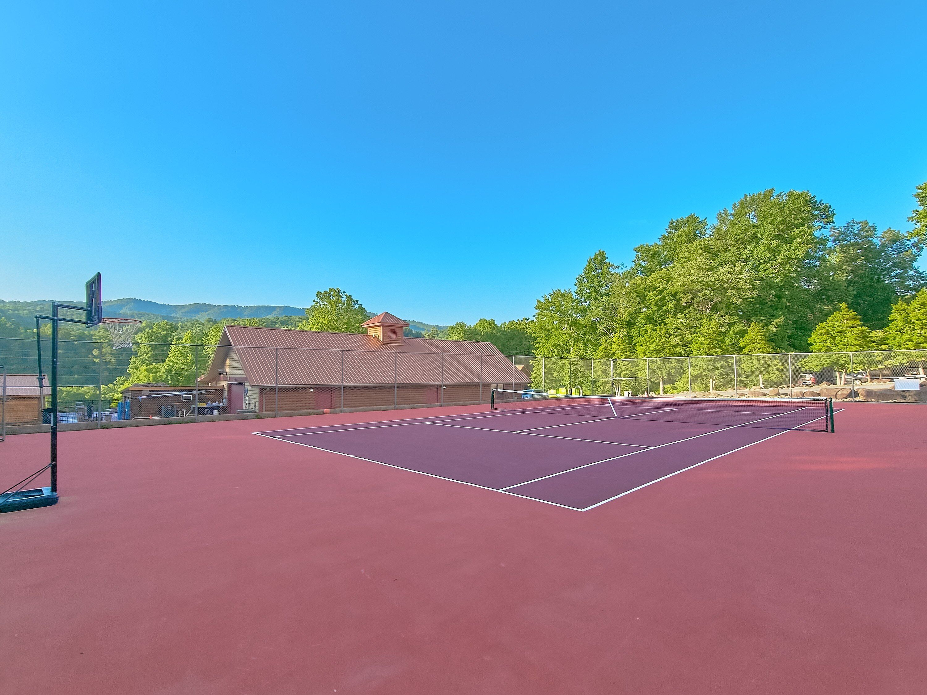 An outdoor tennis court with a net, surrounded by a fence, and a building with a sloped roof in the background.
