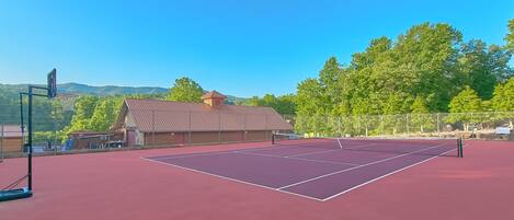 Una cancha de tenis al aire libre con red, rodeada por una valla, y un edificio con techo inclinado al fondo.