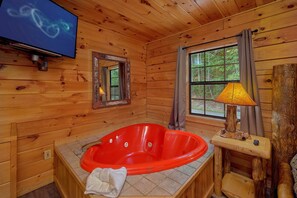 A wooden cabin interior with a red jacuzzi tub, a television mounted on the wall, and a window with a view of greenery. - One More Night | Smoky Mtn Views, Hot Tub, Near Dollywood, Game Room + Free Attraction Tickets (Pigeon Forge)