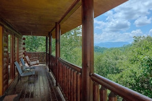 A wooden cabin with a covered porch, outdoor seating, and a scenic view of a forested mountain range. - One More Night | Smoky Mtn Views, Hot Tub, Near Dollywood, Game Room + Free Attraction Tickets (Pigeon Forge)