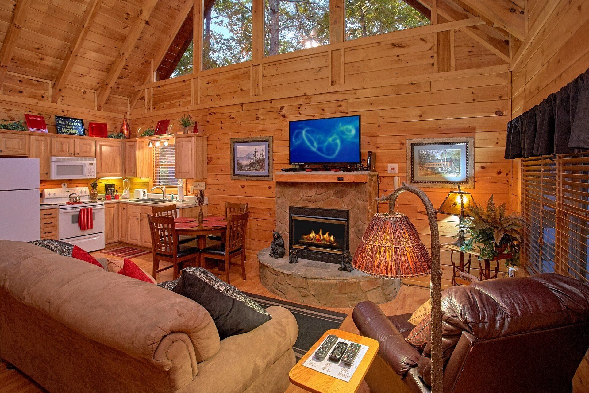 A cozy cabin interior with a stone fireplace, wooden walls, a dining area, and a kitchen.