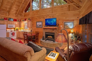 A cozy cabin interior with a stone fireplace, wooden walls, a dining area, and a kitchen. - Our Happy Place | Hot Tub, Near Dollywood, Game Room, Fireplace + Free Attraction Tickets (Pigeon Forge)