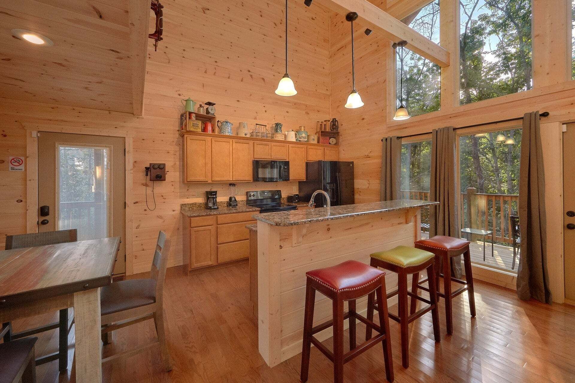A wooden cabin interior with a kitchen, dining area, and large windows.