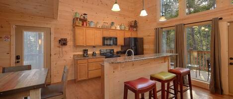 A wooden cabin interior with a kitchen, dining area, and large windows.