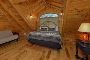A bedroom with a sloped ceiling, a bed with a patterned bedspread, a nightstand with a lamp, and a window with blinds. - Grand Mountain Lodge | Sleeps 26, Theater, Mtn Views, Hot Tub, Game Room + Free Attraction Tickets (Pigeon Forge)