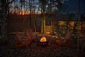 Un foyer extérieur avec deux fauteuils Adirondack, situé dans une zone boisée, avec une terrasse en bois et un bâtiment en arrière-plan.