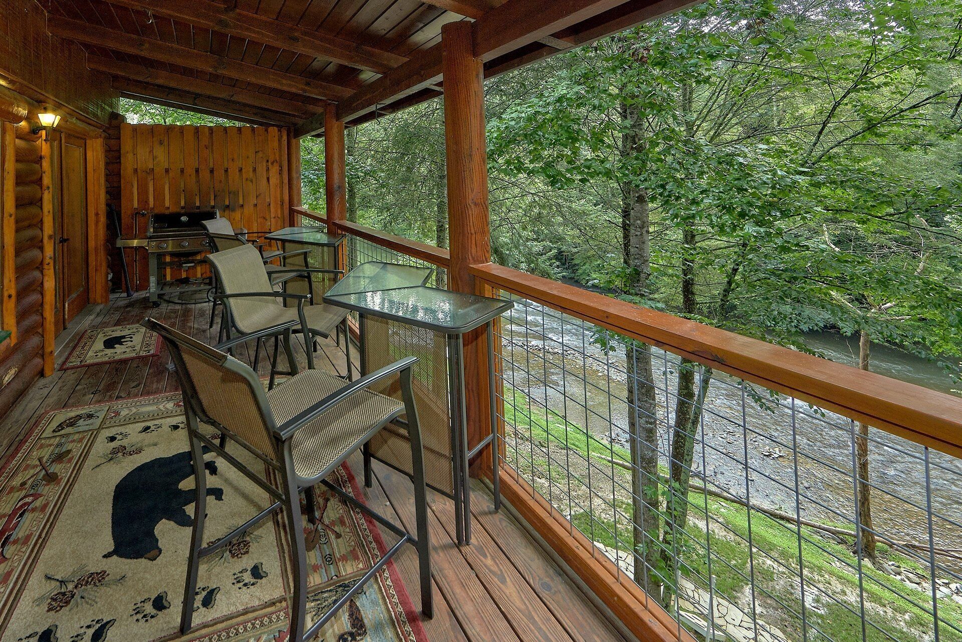 A wooden deck with a glass table and chairs, overlooking a river and forest.