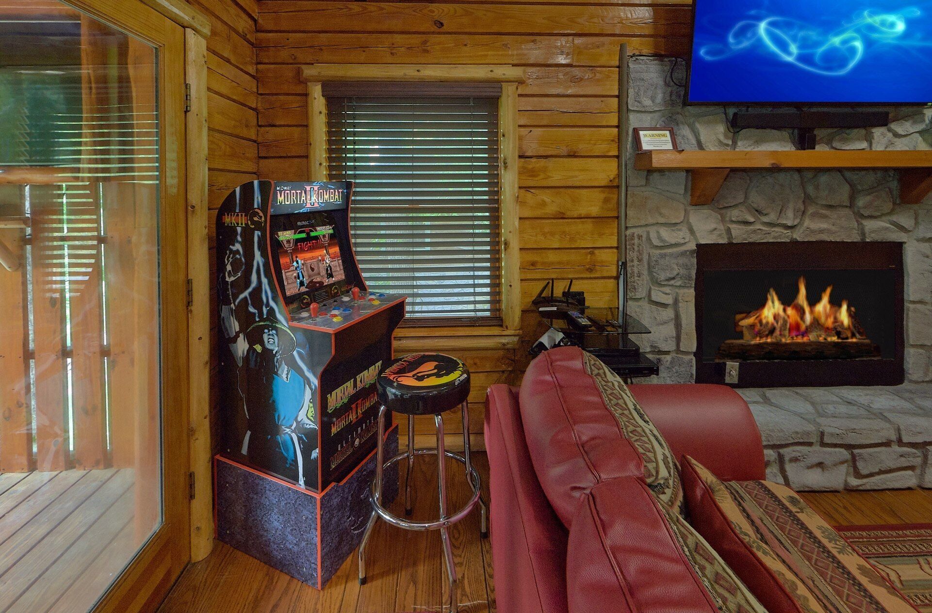 Un intérieur de chalet chaleureux, avec un revêtement mural en bois, une cheminée, un canapé en cuir rouge et une borne d’arcade du jeu Mortal Kombat.