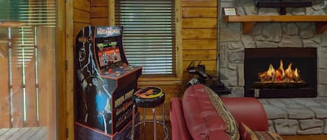 A cozy cabin interior with a wooden interior, a fireplace, a red leather couch, and a Mortal Kombat arcade game.
