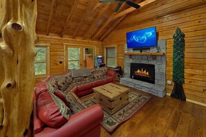A log cabin interior with a stone fireplace, a television mounted above it, and a red leather sofa. - River Edge | Hot Tub, Game Room, Sleeps 10, Fire Pit, Pet Friendly + Free Attraction Tickets (Sevierville)