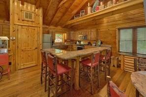 A log cabin interior with a kitchen area featuring wooden cabinets, a refrigerator, a microwave, and a stove. There is a bar area with stools and a wooden table. - River Edge | Hot Tub, Game Room, Sleeps 10, Fire Pit, Pet Friendly + Free Attraction Tickets (Sevierville)