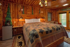 A wooden cabin-style bedroom with a large bed, two bedside lamps, a refrigerator, and a window with blinds. - River Edge | Hot Tub, Game Room, Sleeps 10, Fire Pit, Pet Friendly + Free Attraction Tickets (Sevierville)
