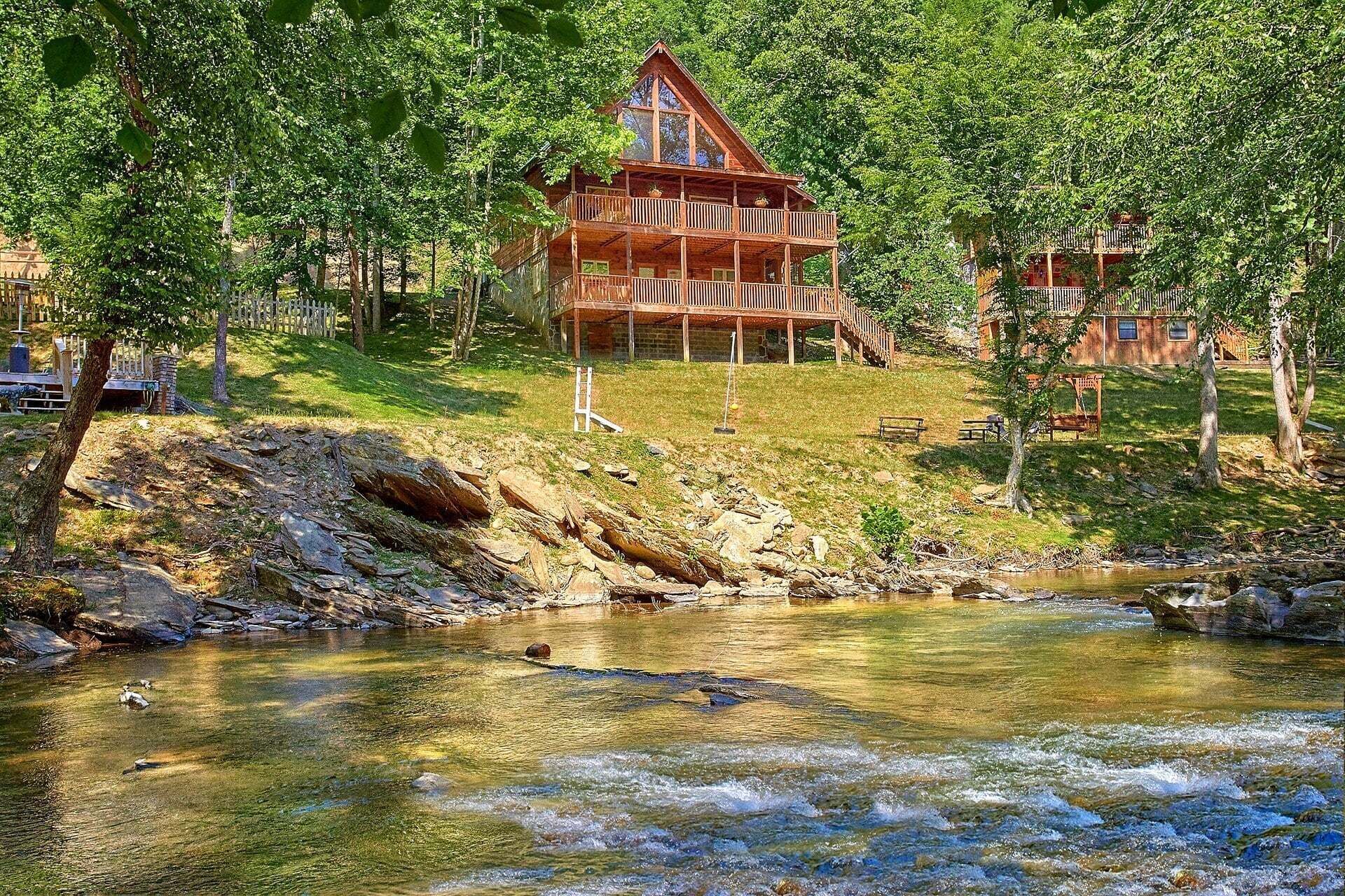 A wooden cabin with a balcony, surrounded by trees and a clear stream.