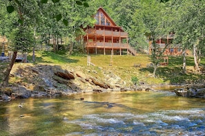 A wooden cabin with a balcony, surrounded by trees and a clear stream. - River Pleasures | Hot Tub, Game Room, Sleeps 10, Fire Pit, Pet Friendly + Free Attraction Tickets (Sevierville)