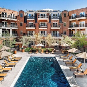 Un espace aménagé au bord de la piscine, avec des chaises longues et des parasols, entouré de bâtiments en briques dotés de balcons.