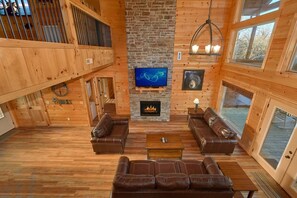 Ein rustikaler Innenraum einer Blockhütte mit Kamin, Ledersofas, Fernseher und Steinwand.