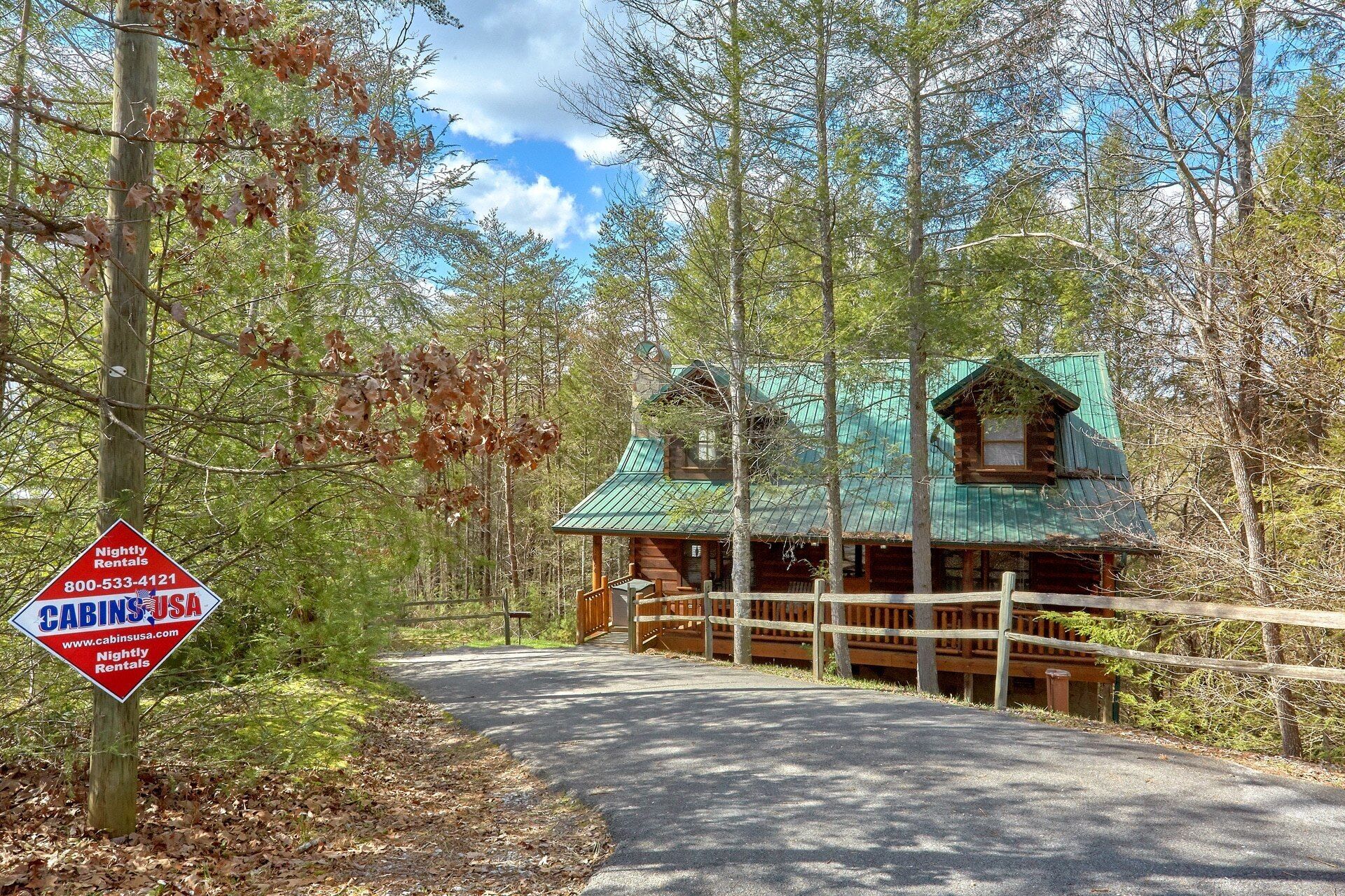 A rustic cabin with a metal roof, surrounded by a wooden fence and trees. A sign indicates it offers nightly rentals.