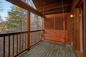 A wooden porch with a swing seat, a railing, and a wall-mounted light fixture. - Saw'n Logs | Hot Tub, Pigeon Forge, Fireplace + Free Attraction Tickets (Pigeon Forge)