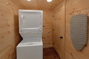 A compact room with a television mounted on a washing machine, wooden walls, and a patterned wall decoration. - Scenic Mountain Pool | Private Pool, Hot Tub, Near Dollywood, Game Room + Free Attraction Tickets (Pigeon Forge)